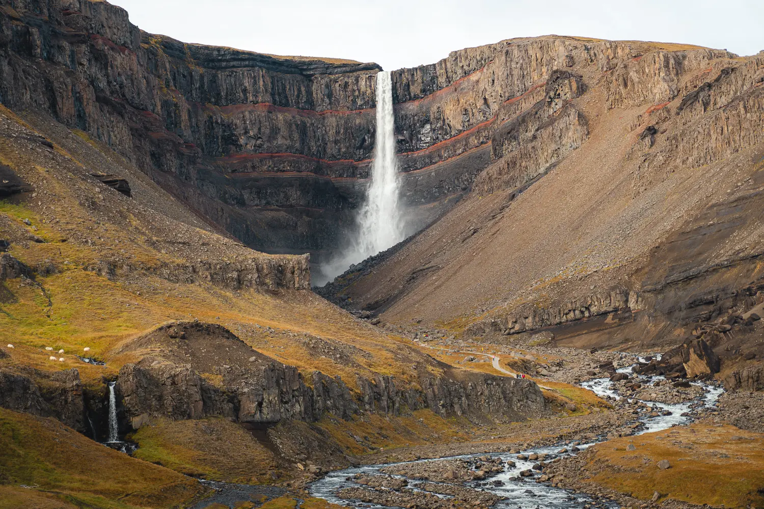 Cascada Hengifoss vista desde abajo, con sus altas paredes de roca y franjas rojizas en el este de Islandia.