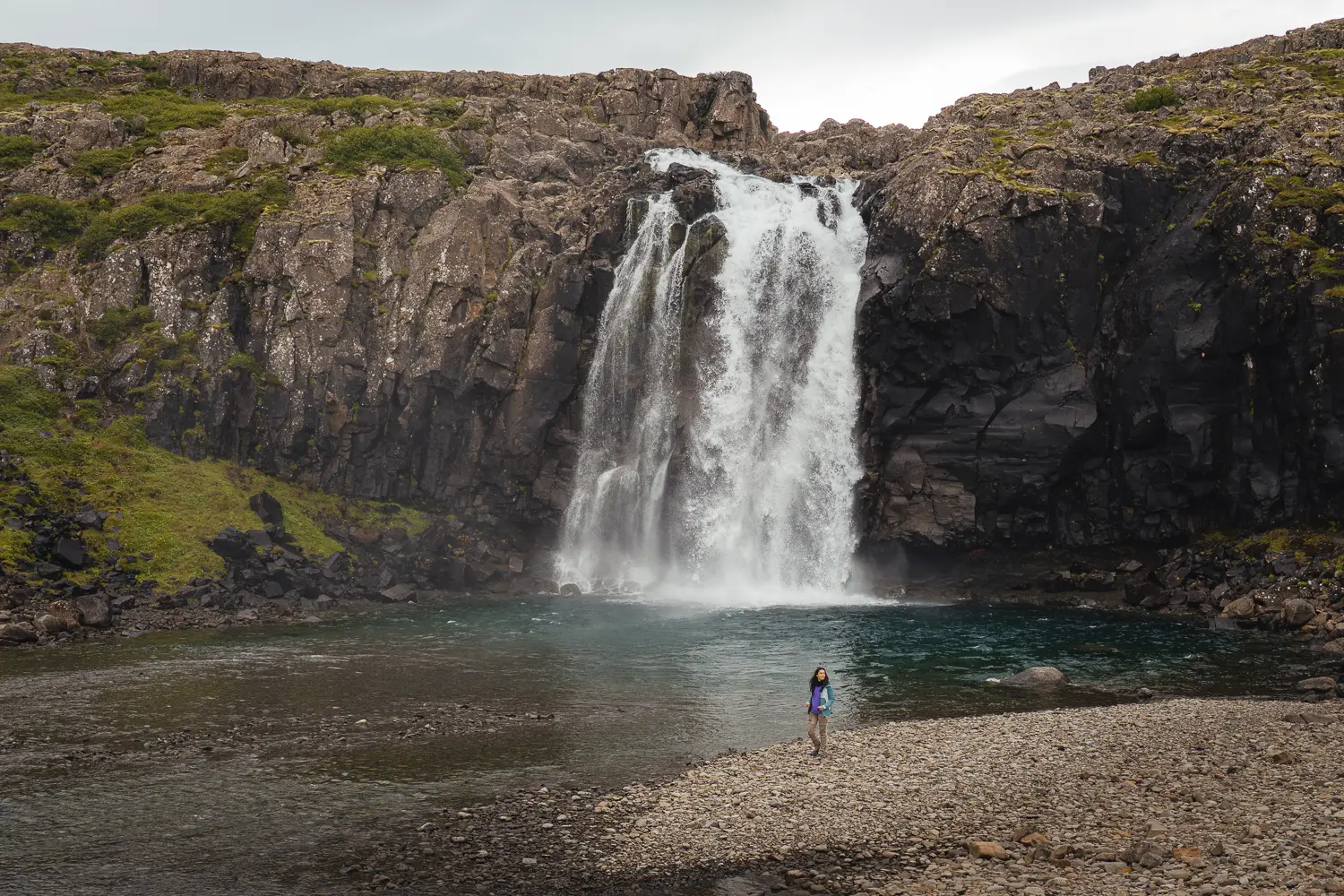 Cascada con un ancho salto de agua que cae por la roca a una poza en color azulón. Se ve las dimensiones de está porque Silvia está frente a la cascada y se la ve extremadamente pequeña.