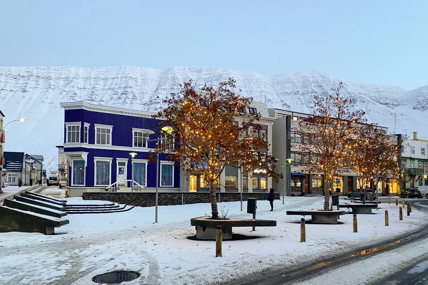 Calle de Ísafjörður al caer la noche con luces decorativas en los árboles. Hay un edificio clásico islandés en azul, y tanto la calle como la montaña de fondo están nevadas