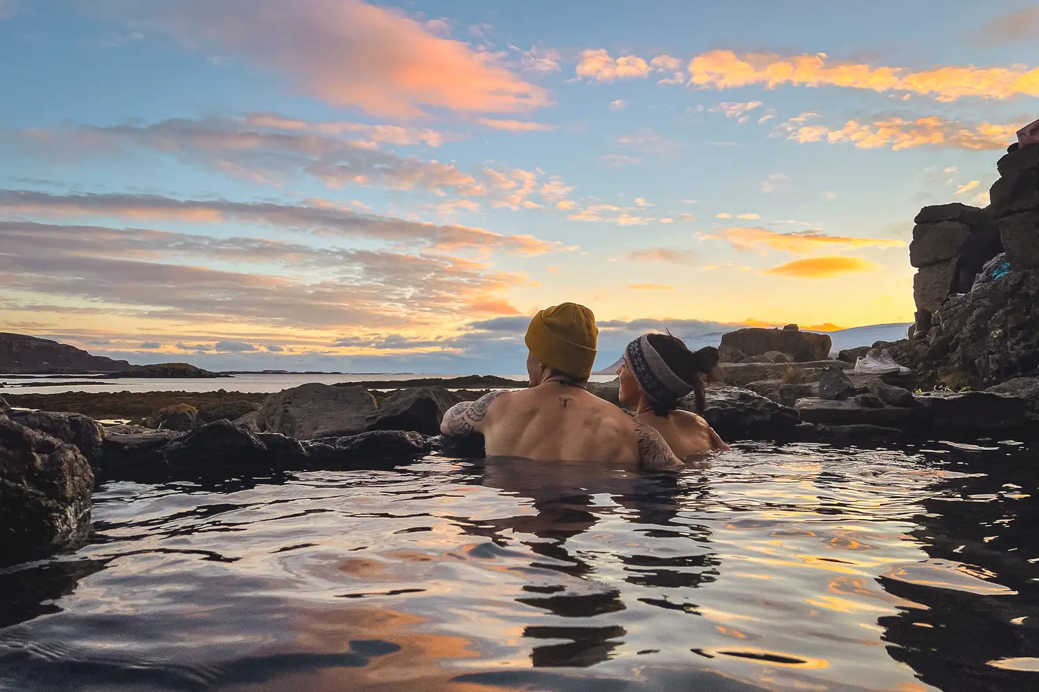 David y Silvia viendo el atardecer, con el cielo en tonos rosas y naranjas desde la poza termal natural Hellulaug frente al océano.