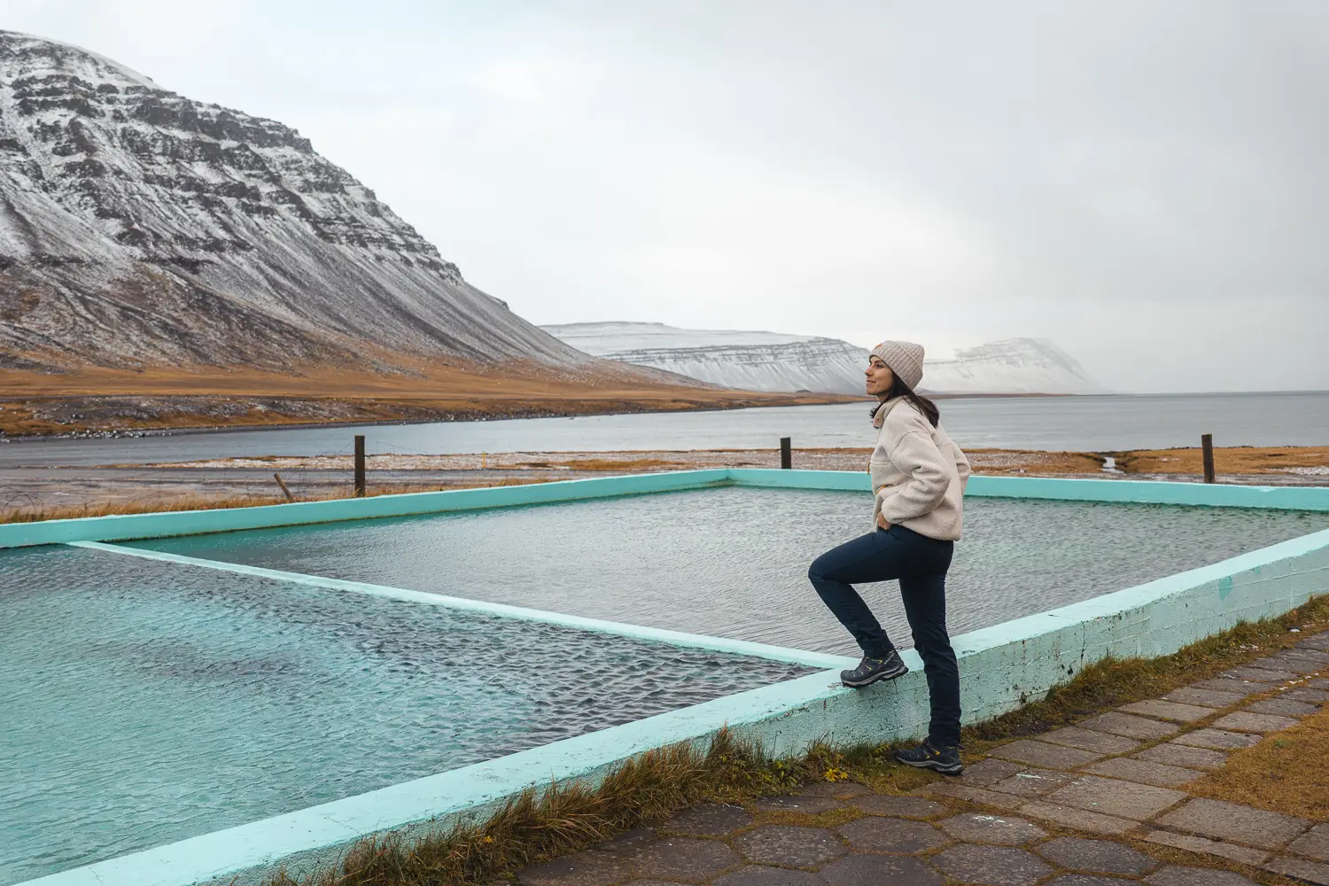 Doble piscina con aguas termales Reykjafjarðarlaug frente al fiordo. Las montañas del fondo están seminevadas.