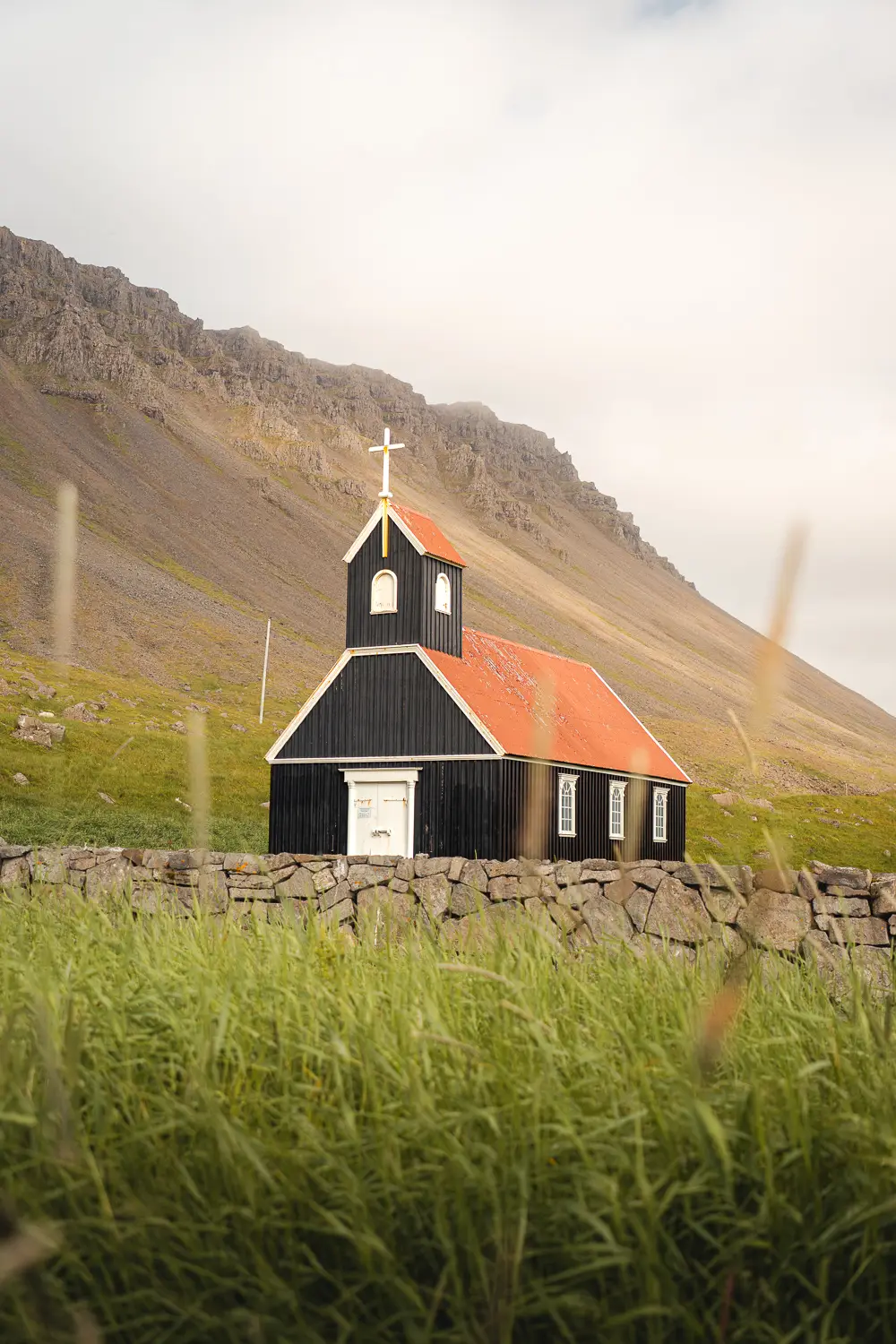 Pequeña iglesia de color negro con el tejado naranja con una montaña de fondo y rodeada de un pequeño muro de piedra. El césped verde rodea el templo.