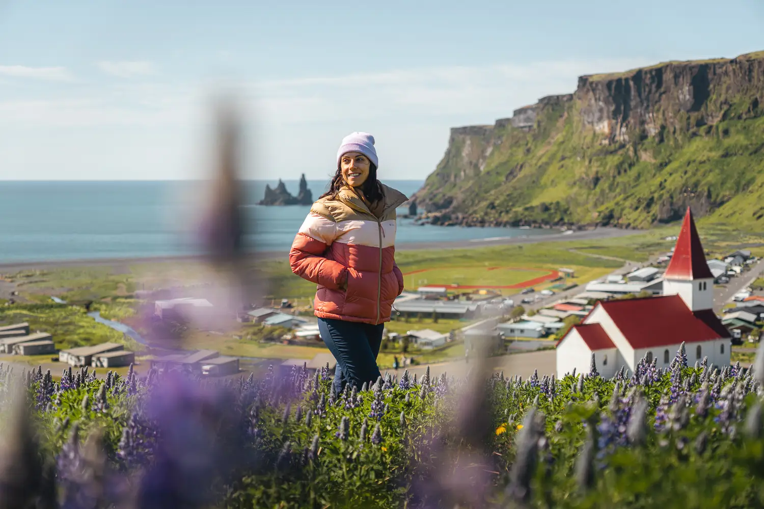 Silvia paseando entre lupines con la vista a la iglesia, playa y ciudad de Vík de fondo.