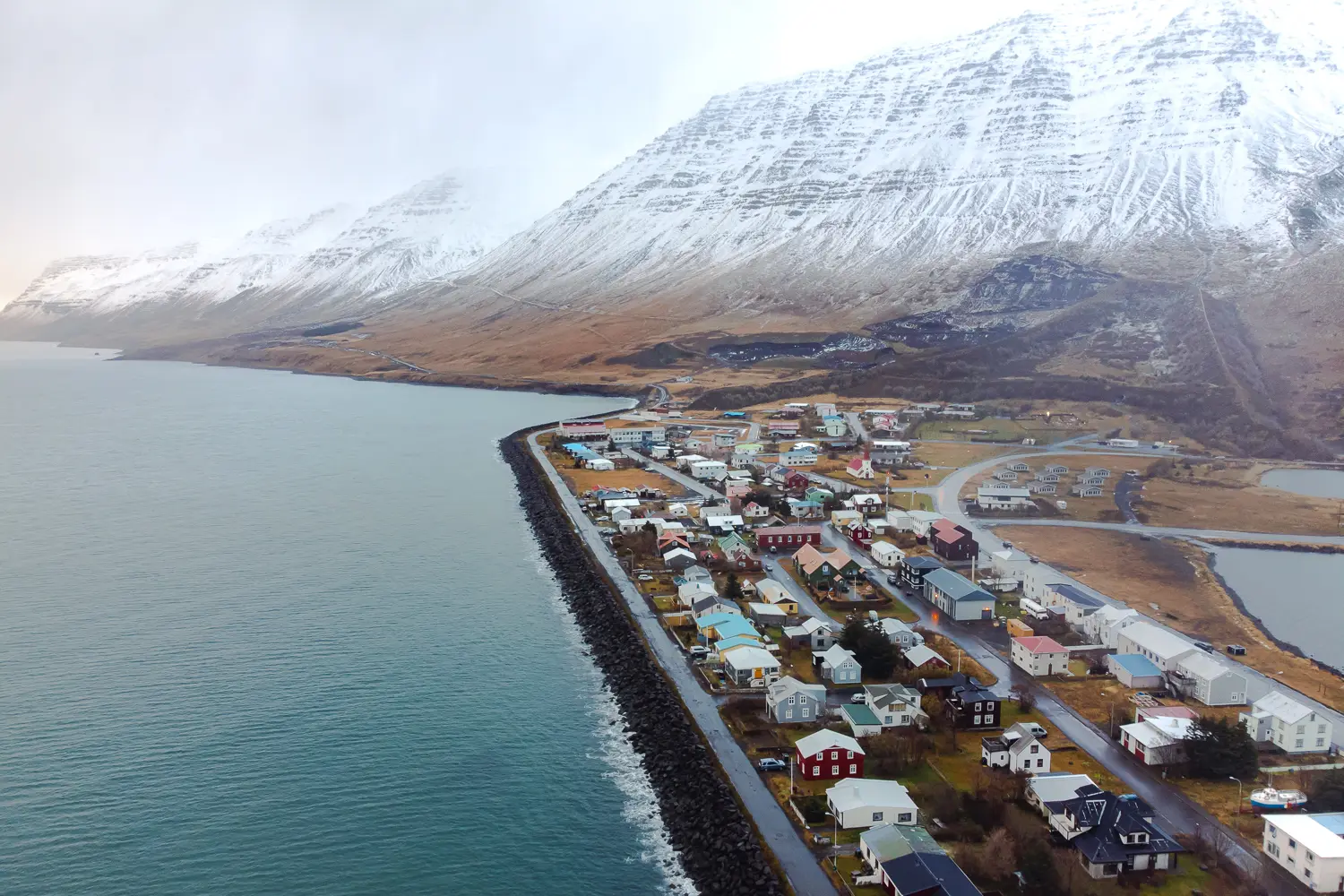 vista de dron del pueblo de Flateyri con sus casas de colores y rodeado de mar con las montañas del fiordo nevadas.