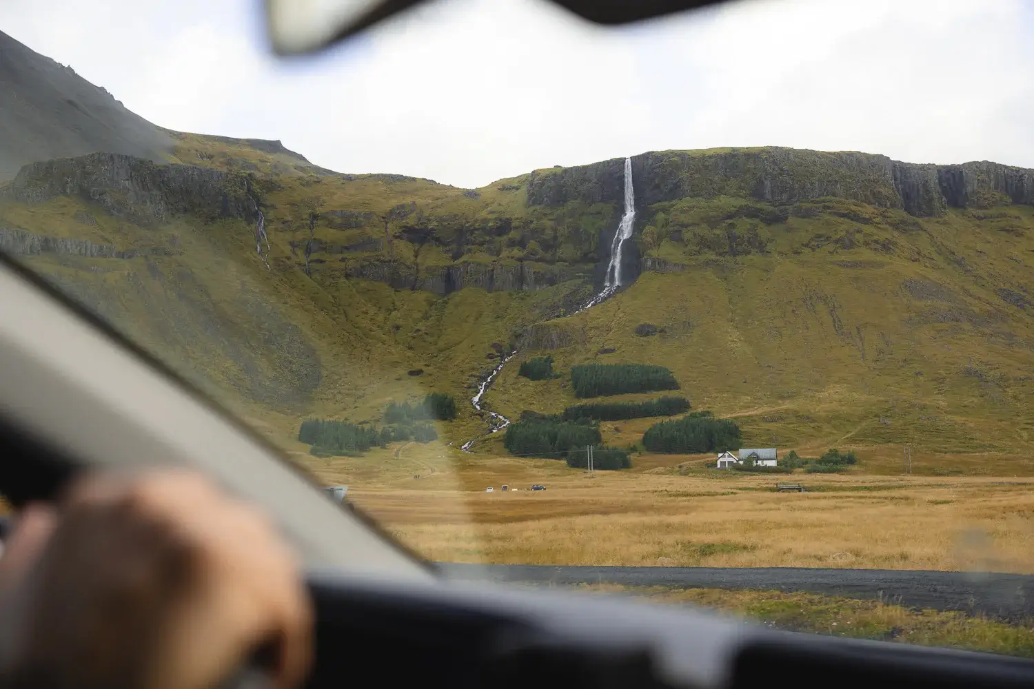 Cascada junto a la carretera en un paisaje rural de Islandia con la vista desde el interior del coche de alquiler